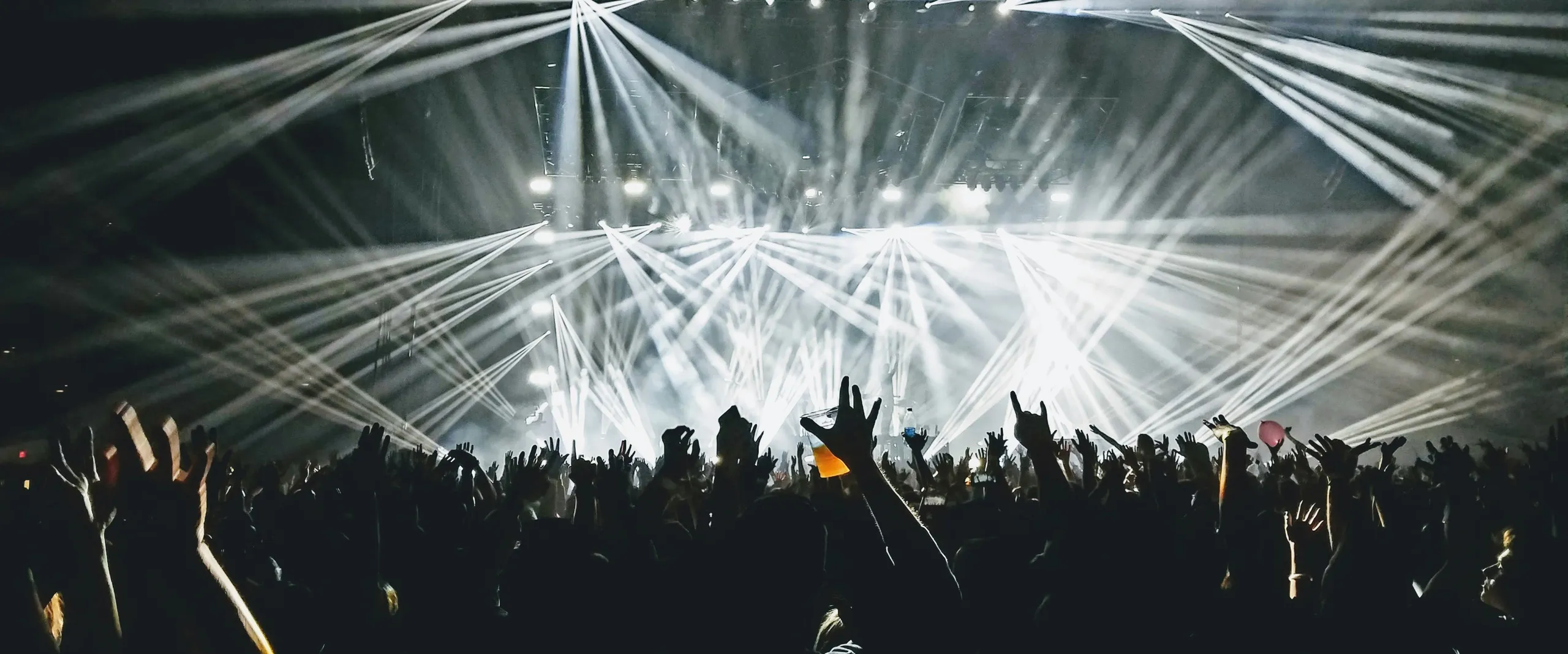 Concert crowd with raised hands in front of a stage lit by beams of white light